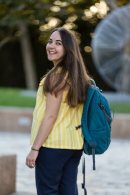 Viven standing in a park wearing a yellow blouse and carrying a turquoise backpack.